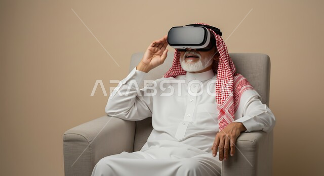 استخدام جهاز تقني حديث، المتعة والتسلية بالمشاهدة عبر نظارة الواقع الافتراضي، إيماءات الاندماج والتركيز، بورتريه لرجل عربي خليجي سعودي كبير بالسن يرتدي الثوب التقليدي والشماغ يجلس على كرسي مريح ينظر من خلال نظارات ثلاثية الأبعاد، محاكاة العالم الخيالي، خلفيه بيج