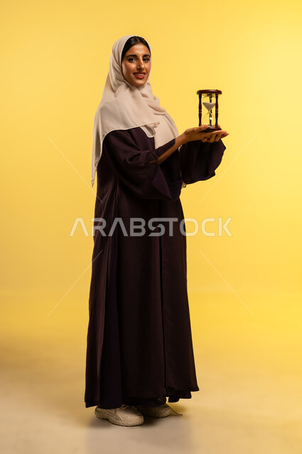 تحديد مواعيد العمل وتنظيم الجدول الزمني، مفهوم إدارة الأعمال والأوقات، شابة سعودية تنظر الى الكاميرا بتعابير السرور، بورتريه لامرأة عربية خليجية عمانية ترتدي الحجاب والعباءة تحمل الساعة الرملية الزجاجية (البنكام) بايماءات أهميه مراقبة الوقت, الجسم بالطول الكامل، خلفية صفراء