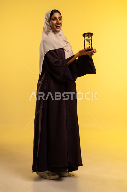 مفهوم إدارة الأعمال والأوقات، شابة سعودية تنظر الى الكاميرا بتعابير السرور، بورتريه لامرأة عربية خليجية عمانية ترتدي الحجاب والعباءة تحمل الساعة الرملية الزجاجية (البنكام) بايماءات أهميه مراقبة الوقت, تحديد مواعيد العمل وتنظيم الجدول الزمني، الجسم بالطول الكامل، خلفية صفراء
