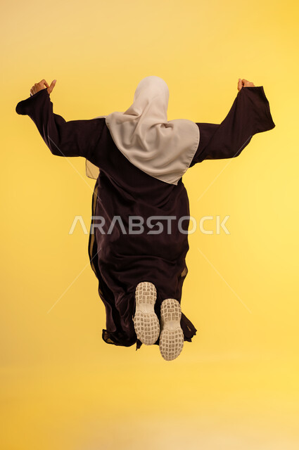 Elegance and interest in appearance, concept of femininity and softness, young Saudi woman jumping high with gestures of joy and happiness, portrait from behind of an Arab Gulf Omani woman wearing hijab and abaya raising her hands with expressions of happiness, full body, yellow background
