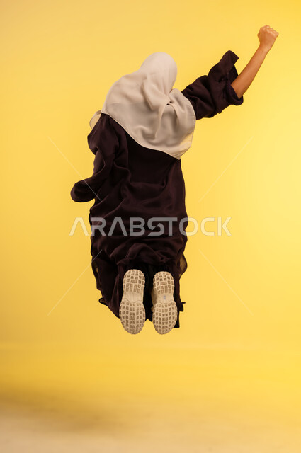 The concept of femininity and softness, interest in external appearance, a young Saudi woman jumping high with gestures of joy and happiness, a portrait from the back of an Arab Gulf Omani woman wearing a hijab and an abaya raising her hand with expressions of happiness, full-length body, yellow background