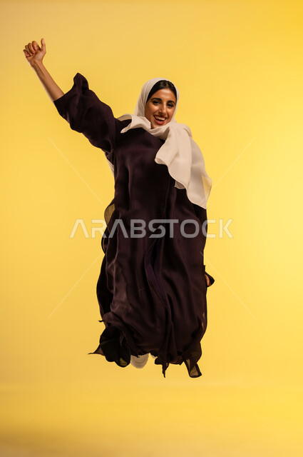 الاهتمام بالمظهر الخارجي، مفهوم الأنوثة والنعومة، شابة سعودية تقفز عاليا بايماءات الفرح والسرور، بورتريه لامرأة عربية خليجية عمانية ترتدي الحجاب والعباءة ترفع يدها وتنظر إلى الكاميرا بتعابير السعادة, الجسم بالطول الكامل، خلفية صفراء