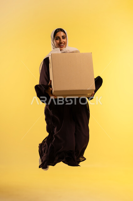 التوصيل والشحن ذات الجودة العالية، طلب المنتجات والتسوق عبر الإنترنت، بورتريه لامرأة عربية خليجية عمانية محجبة ترتدي عباءة بنية تحمل بيدها صندوق كرتوني، شابة سعودية تقفز عاليا بتعابير الفرح، استلام الطلبات الاونلاين من باب المنزل، الطول الكامل للجسم، خلفيه صفراء