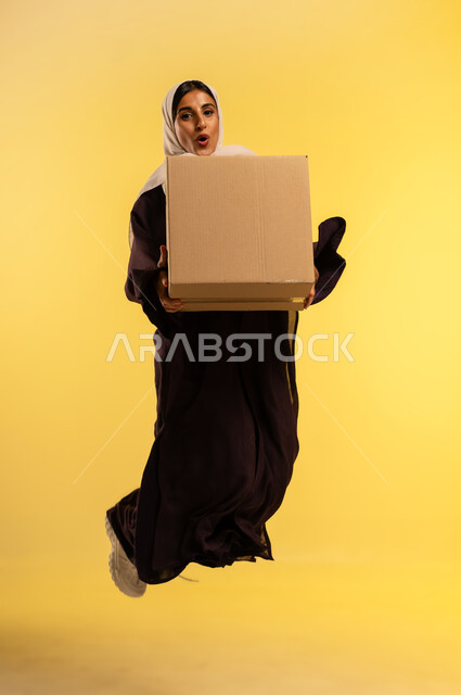 طلب المنتجات والتسوق عبر الإنترنت، التوصيل والشحن ذات الجودة العالية، بورتريه لامرأة عربية خليجية عمانية محجبة ترتدي عباءة بنية تحمل بيدها صندوق كرتوني، شابة سعودية تقفز عاليا بتعابير الفرح، استلام الطلبات الاونلاين من باب المنزل، الطول الكامل للجسم، خلفيه صفراء