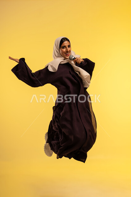 مفهوم الفن وتأثيره الايجابي على المرأة، تقديم العروض الفنيه الغنائيه والفقرات الترفيهية، بدء التدريب الصوتي، بورتريه لامرأة عربية خليجية عمانية محجبة ترتدي عباءة بنية تمسك بيدها المايكروفون، شابة سعودية تقفز عاليا بتعابير الفرح، الطول الكامل للجسم، خلفيه صفراء
