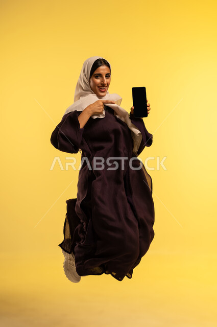 الدردشة مع الاصدقاء عبر الجوال، استخدام جهاز تقني حديث، بورتريه لامرأة عربية خليجية عمانية ترتدي العباءة البنية والحجاب تقوم بعرض شاشة سوداء فارغة عبر الهاتف المحمول، شابة سعودية تقفز عاليا بإيماءات الفرح، الجسم بالطول الكامل، خلفية صفراء