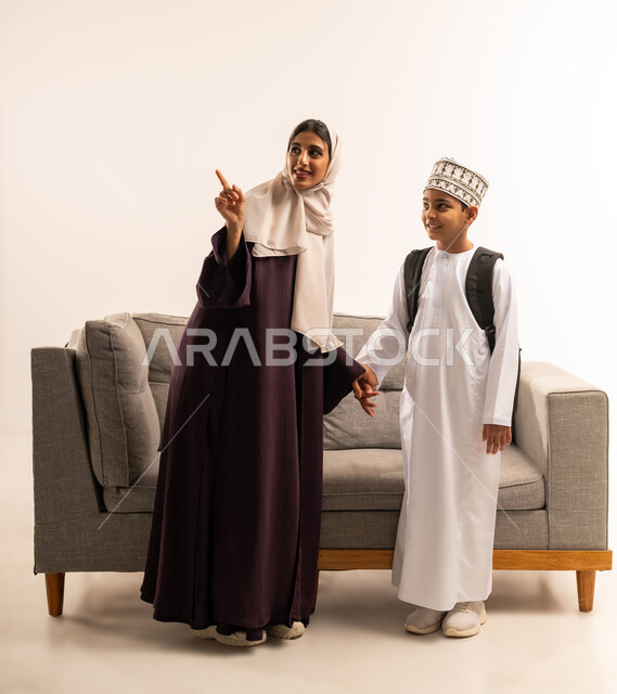الاجتهاد والمثابرة للوصول الى المراتب العليا، مفهوم التعلم في المدارس المتطورة في سلطنة عمان، الاستعداد لبدء عام دراسي جديد، بورتريه لأم عربية خليجية عمانية محجبة ترتدي عباءة بنية تقوم بمساعدة ابنها بالتجهز للذهاب الى المدرسة، الجسم بالطول الكامل، خلفية بيضاء