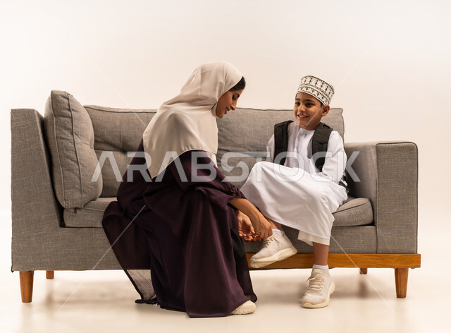 مفهوم التعلم في المدارس المتطورة في سلطنة عمان، الاستعداد لبدء عام دراسي جديد، المثابرة والاجتهاد للوصول الى المراتب العليا، بورتريه لأم عربية خليجية عمانية محجبة ترتدي عباءة بنية تقوم بمساعدة ابنها بالتجهز للذهاب الى المدرسة، خلفية بيضاء