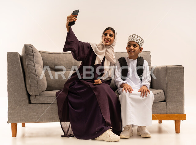 التقاط صور سيلفي عبر الهاتف المحمول، الاستعداد لبدء عام دراسي جديد، الوصول الى المراتب العليا بالاجتهاد والمثابرة، التعلم في المدارس المتطورة في سلطنة عمان، بورتريه لأم عربية خليجية عمانية محجبة ترتدي عباءة بنية تجلس على اريكة رمادية مع ابنها، خلفية بيضاء