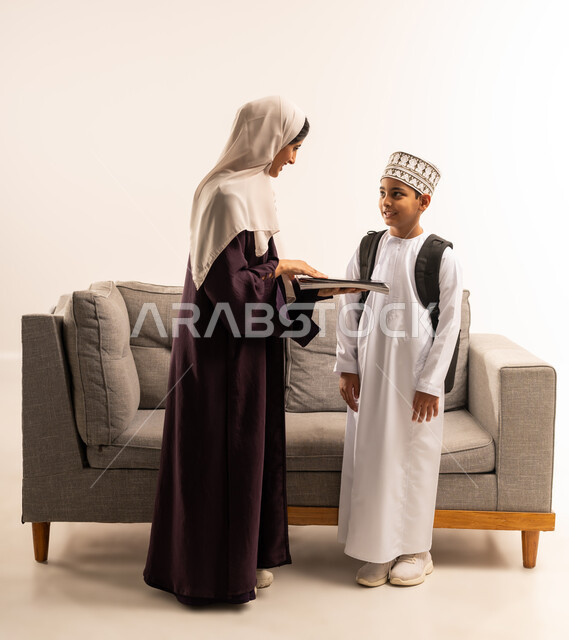 الاستعداد لبدء عام دراسي جديد، المثابرة والاجتهاد للوصول الى المراتب العليا، مفهوم التعلم في المدارس المتطورة في سلطنة عمان، بورتريه لأم عربية خليجية عمانية محجبة ترتدي عباءة بنية تقوم بمساعدة ابنها بالتجهز للذهاب الى المدرسة، الجسم بالطول الكامل، خلفية بيضاء