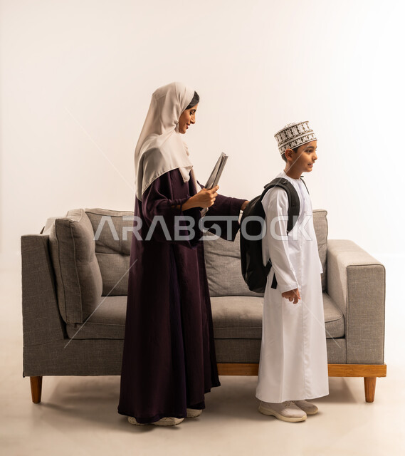 المثابرة والاجتهاد للوصول الى المراتب العليا، مفهوم التعلم في المدارس المتطورة في سلطنة عمان، الاستعداد لبدء عام دراسي جديد، بورتريه لأم عربية خليجية عمانية محجبة ترتدي عباءة بنية تقوم بمساعدة ابنها بالتجهز للذهاب الى المدرسة، الجسم بالطول الكامل، خلفية بيضاء
