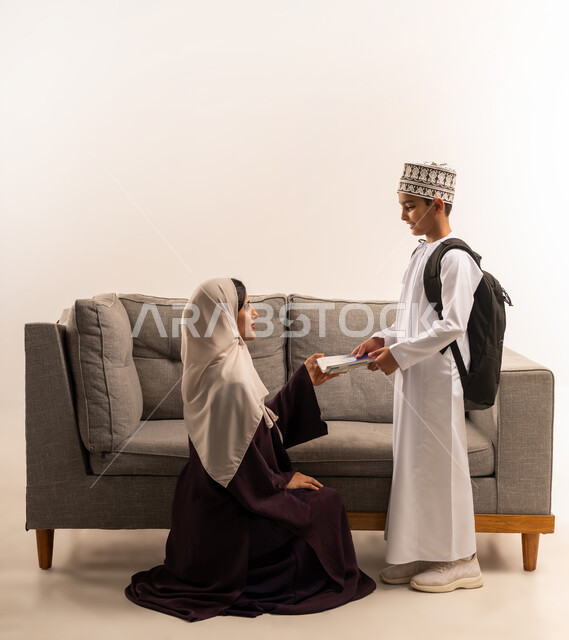 The concept of hard work and perseverance to reach the highest ranks, preparing to start a new school year, learning in advanced schools in the Sultanate of Oman, portrait of a veiled Gulf Arab Omani mother wearing a brown abaya helping her son get ready to go to school, full body, white background