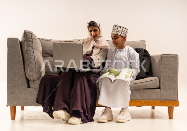 تشجيع وتطوير الابناء بحل الواجبات المدرسيه المنزليه، تنميه قدرات الاطفال بالتعلم، بورتريه لأم عربية خليجية عمانيه محجبة تجلس على اريكة رمادية وتساعد ابنها في التعليم والقراءه، الانشطة التعليمية الالكترونيه وانجاز الواجبات عن بعد باستخدام جهاز الحاسوب المحمول، خلفية بيضاء