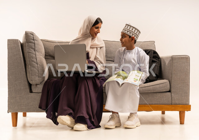 تنميه قدرات الاطفال بالتعلم، تشجيع وتطوير الابناء بحل الواجبات المدرسيه المنزليه، بورتريه لأم عربية خليجية عمانيه محجبة تجلس على اريكة رمادية وتساعد ابنها في التعليم والقراءه، الانشطة التعليمية الالكترونيه وانجاز الواجبات عن بعد باستخدام جهاز الحاسوب المحمول، خلفية بيضاء