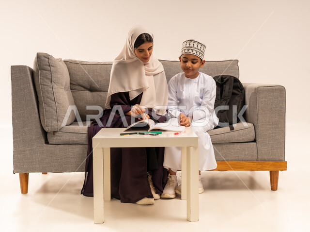 الأنشطة التعليمية وتنميه قدرات الاطفال بالتعلم، دعم وتشجيع الابناء لحل الدروس والواجبات المدرسيه المنزليه، بورتريه لأم عربية خليجية عمانيه محجبة تجلس على اريكة رمادية وتساعد ابنها في التعليم والقراءه، الاهتمام بتعليم الأبناء، خلفية بيضاء
