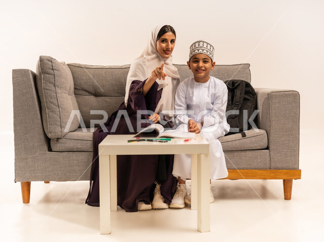 دعم وتشجيع الابناء لحل الدروس والواجبات المدرسيه المنزليه، الأنشطة التعليمية وتنميه قدرات الاطفال بالتعلم، بورتريه لأم عربية خليجية عمانيه محجبة تجلس على اريكة رمادية وتساعد ابنها في التعليم والقراءه، الاهتمام بتعليم الأبناء، خلفية بيضاء