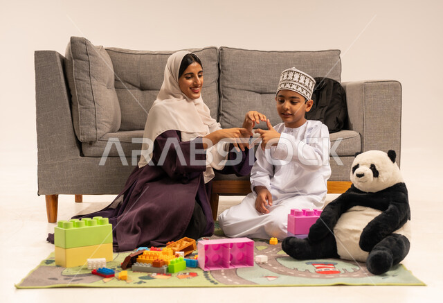 قضاء وقت ممتع مع الوالدة بايماءات السعادة والسرور ، الاهتمام بالأبناء ودعمهم، مفهوم الأمومة والطفولة، رعاية وتشجيع وتطوير مهارات الأطفال، الاستمتاع بتركيب المكعبات، بورتريه لام عربية خليجية عمانية محجبة ترتدي عباءة بنية تلعب مع ابنها، خلفية بيضاء