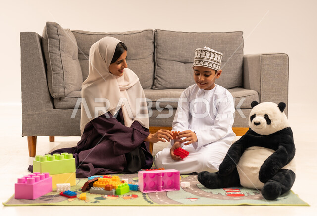 Caring for, encouraging and developing children's skills, gestures of happiness and pleasure in spending quality time with the mother, enjoying assembling blocks, supporting and caring for children, the concept of motherhood and childhood, portrait of a veiled Gulf Arab Omani mother wearing a brown abaya playing with her son, white background
