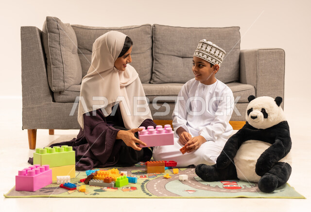 تشجيع ورعاية وتطوير مهارات الأطفال، ايماءات السعادة والسرور بقضاء وقت ممتع مع الوالدة، الاستمتاع بتركيب المكعبات، دعم الأبناء والاهتمام بهم، مفهوم الأمومة والطفولة، بورتريه لام عربية خليجية عمانية محجبة ترتدي عباءة بنية تلعب مع ابنها، خلفية بيضاء