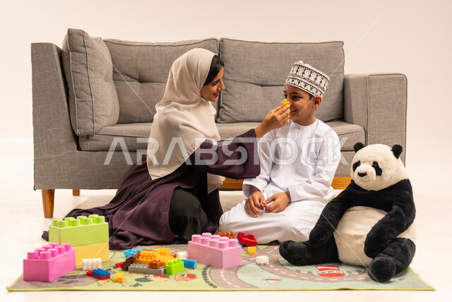 ايماءات السعادة والسرور بقضاء وقت ممتع مع الوالدة، الاستمتاع بتركيب المكعبات، دعم الأبناء والاهتمام بهم، تشجيع ورعاية وتطوير مهارات الأطفال، مفهوم الأمومة والطفولة، بورتريه لام عربية خليجية عمانية محجبة ترتدي عباءة بنية تلعب مع ابنها، خلفية بيضاء
