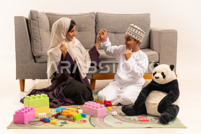 تعابير السعادة والسرور بقضاء وقت ممتع مع الوالدة، الاستمتاع بتركيب المكعبات، دعم الأبناء والاهتمام بهم، تشجيع ورعاية وتطوير مهارات الأطفال، مفهوم الأمومة والطفولة، بورتريه لام عربية خليجية عمانية محجبة ترتدي عباءة بنية تلعب مع ابنها، خلفية بيضاء