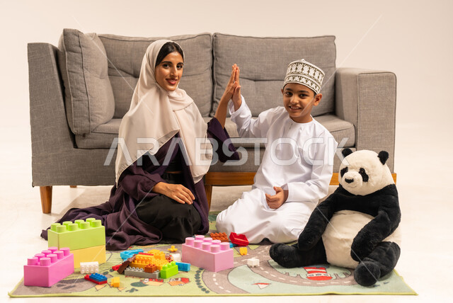 الاستمتاع بتركيب المكعبات، دعم الأبناء والاهتمام بهم، تشجيع ورعاية وتطوير مهارات الأطفال، مفهوم الأمومة والطفولة، تعابير السعادة والسرور بقضاء وقت ممتع مع الوالدة، بورتريه لام عربية خليجية عمانية محجبة ترتدي عباءة بنية تلعب مع ابنها، خلفية بيضاء