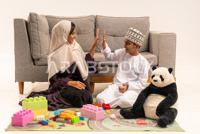Supporting and caring for children, encouraging, caring for and developing children's skills, the concept of motherhood and childhood, expressions of happiness and pleasure in spending a fun time with the mother, enjoying assembling cubes, portrait of a veiled Gulf Arab Omani mother wearing a brown abaya playing with her son, white background