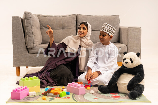 Caring for and supporting children, encouraging, nurturing and developing children's skills, the concept of motherhood and childhood, expressions of happiness and pleasure in spending a fun time with the mother, enjoying assembling cubes, portrait of a veiled Gulf Arab Omani mother wearing a brown abaya playing with her son, white background