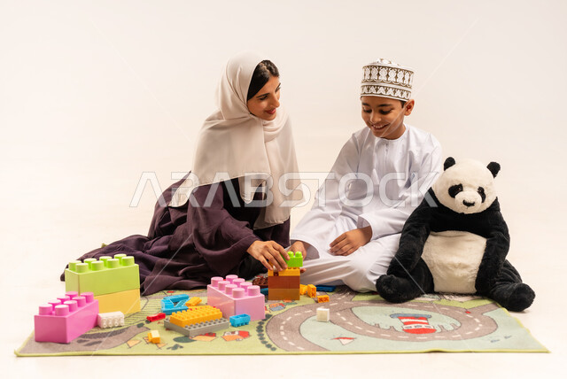Encouraging, nurturing and developing children's skills, the concept of motherhood and childhood, expressions of happiness and pleasure in spending a fun time with the mother, enjoying assembling cubes, portrait of a veiled Gulf Arab Omani mother wearing a brown abaya playing with her son, caring for and supporting children, white background