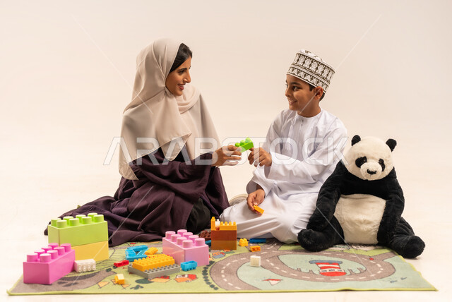 رعاية وتشجيع وتطوير مهارات الأطفال، مفهوم الأمومة والطفولة، تعابير السعادة والسرور بقضاء وقت ممتع مع الوالدة، الاستمتاع بتركيب المكعبات، بورتريه لام عربية خليجية عمانية محجبة ترتدي عباءة بنية تلعب مع ابنها، الاهتمام بالأبناء ودعمهم، خلفية بيضاء
