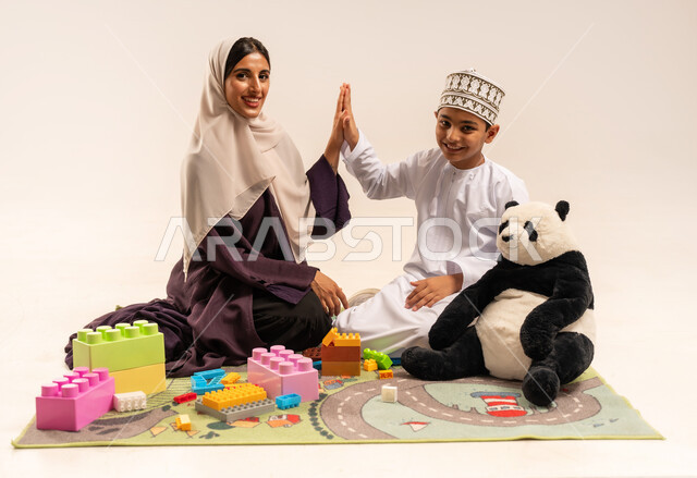 الاستمتاع بتركيب المكعبات، بورتريه لام عربية خليجية عمانية محجبة ترتدي عباءة بنية تلعب مع ابنها، ايماءات السعادة والسرور بقضاء وقت ممتع مع الوالدة، مفهوم الأمومة والطفولة، رعاية وتشجيع وتطوير مهارات الأطفال، الاهتمام بالأبناء ودعمهم، خلفية بيضاء