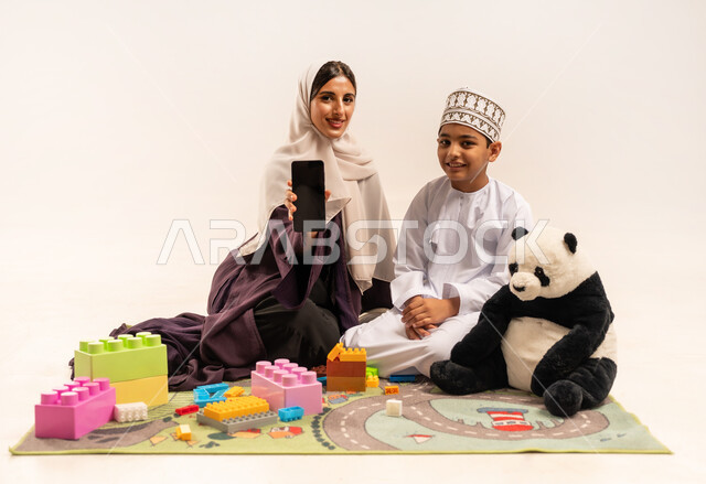 قضاء وقت ممتع مع الوالدة بايماءات السعادة، الاهتمام بالأبناء ودعمهم، مفهوم الأمومة والطفولة، تشجيع وتطوير مهارات الأطفال، الاستمتاع بتركيب المكعبات، بورتريه لام عربية خليجية عمانية محجبة ترتدي عباءة بنية تقوم بعرض شاشة سوداء فارغة عبر الهاتف المحمول، خلفية بيضاء