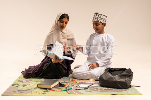 الاهتمام بتعليم الأبناء ودعمهم، بورتريه لام عربية خليجية عمانية محجبة ترتدي عباءة بنية تجلس مع ابنها على حصيرة وتقوم بمساعدة ابنها في حل الوظائف، ولد عماني يرتدي الدشداشة والكمة يقوم بتأدية الواجبات المدرسية، ، الانشطة التعليمية وتنمية القدرات، خلفية بيضاء