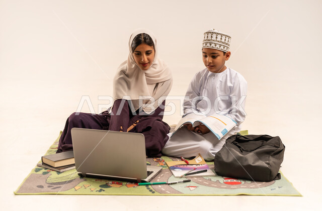 تنميه قدرات الاطفال بالتعلم، انجاز الدروس والواجبات عن بعد باستخدام جهاز الحاسوب المحمول، الانشطة التعليمية الالكترونيه، تطوير وتشجيع الاطفال بحل الواجبات المدرسيه، بورتريه لأم عربية خليجية عمانيه محجبة ترتدي عباءة بنية تساعد طفلها في التعليم والقراءه، خلفية بيضاء