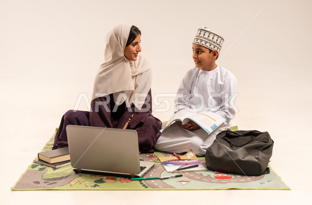 انجاز الدروس والواجبات عن بعد باستخدام جهاز الحاسوب المحمول، الانشطة التعليمية الالكترونيه، تطوير وتشجيع الاطفال بحل الواجبات المدرسيه، تنميه قدرات الاطفال بالتعلم، بورتريه لأم عربية خليجية عمانيه محجبة ترتدي عباءة بنية تساعد طفلها في التعليم والقراءه، خلفية بيضاء
