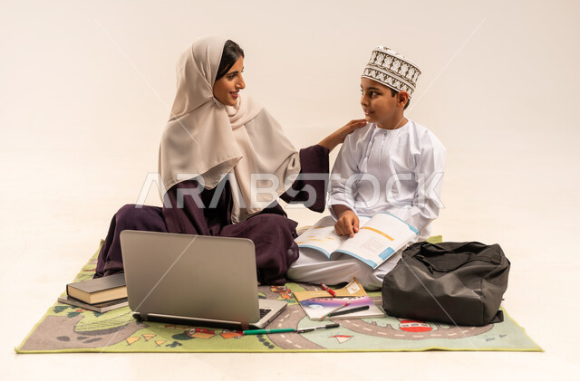 Developing and encouraging children to solve school assignments, developing children's learning abilities, portrait of a veiled Gulf Arab Omani mother wearing an abaya, putting her hand on her son's shoulder, helping children learn and read, electronic educational activities, completing lessons remotely using a laptop, white background