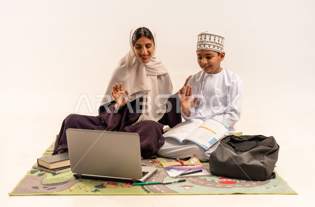 Raising the palm with greeting and peace gestures, encouraging children to do their homework, developing children's abilities, portrait of a veiled Gulf Arab Omani mother wearing an abaya helping her son in education and reading, electronic educational activities, completing lessons remotely using a laptop, white background