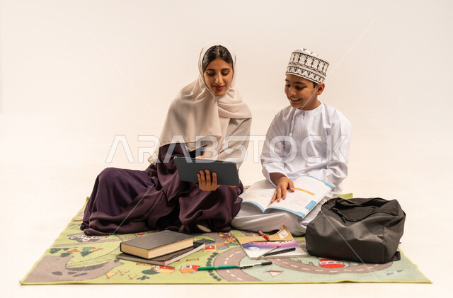 تنميه قدرات الاطفال بالتعلم، تشجيع وتطوير الاطفال بحل الواجبات المدرسيه، بورتريه لأم عربية خليجية عمانيه محجبة ترتدي عباءة بنية تساعد طفلها في التعليم والقراءه، الانشطة التعليمية الالكترونيه، انجاز الدروس والواجبات عن بعد باستخدام جهاز الايباد المحمول، خلفية بيضاء