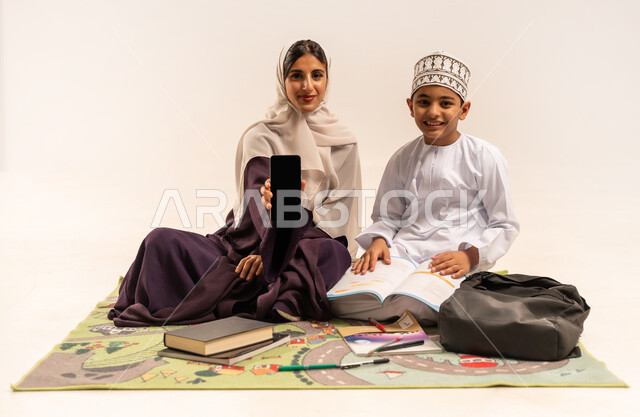 Caring for children's education, developing abilities and educational activities, helping with homework, portrait of a veiled Gulf Arab Omani mother wearing a brown abaya displaying a blank black screen on a mobile phone, Omani boy wearing a dishdasha and a kimono sitting on a mat with his mother, white background