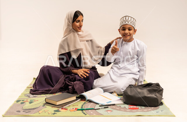الانشطة التعليمية وتنمية القدرات، المساعدة في تأدية الواجبات المدرسية، الاهتمام بتعليم الأبناء، بورتريه لام عربية خليجية عمانية محجبة ترتدي عباءة بنية تجلس مع ابنها على حصيرة وتقوم بتدريسه، ولد عماني يرتدي الدشداشة والكمة يرفع اصبع الابهام بايماءات الاعجاب، خلفية بيضاء