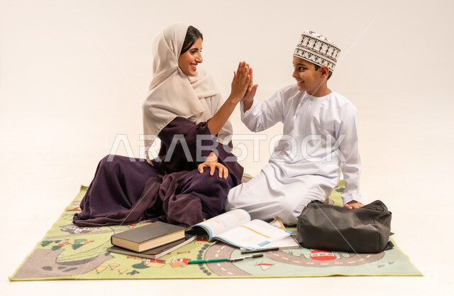 المساعدة في تأدية الواجبات المدرسية، الاهتمام بتعليم الأبناء ودعمهم، بورتريه لام عربية خليجية عمانية محجبة ترتدي عباءة بنية تجلس مع ابنها على حصيرة وتقوم بتدريسه، ولد عماني يرتدي الدشداشة والكمة يضرب كفه بكف والدته، الانشطة التعليمية وتنمية القدرات، خلفية بيضاء
