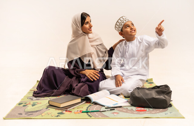الاهتمام بتعليم الأبناء ودعمهم، بورتريه لام عربية خليجية عمانية محجبة ترتدي عباءة بنية تجلس مع ابنها على حصيرة وتقوم بتدريسه، المساعدة في تأدية الواجبات المدرسية، ولد عماني يرتدي الدشداشة والكمة يشير باصبع السبابة للأعلى، الانشطة التعليمية وتنمية القدرات، خلفية بيضاء