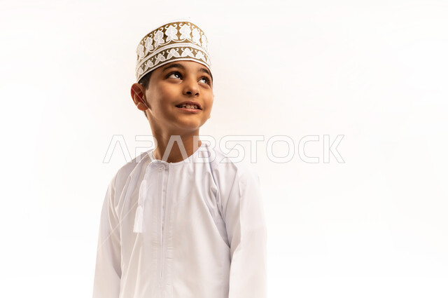 مفهوم الاناقه والاهتمام بالمظهر الخارجي، الوقوف باستقامة وثقه بالنفس، بورتريه مقرب لولد عربي خليجي عماني مبتسم يرتدي الدشداشة والكمة ينظر إلى الأعلى بتعابير السعادة والسرور، خلفية بيضاء