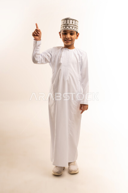 Standing straight and looking at camera, expressions of inquiry and pointing at something, portrait of an Arab Gulf Omani boy wearing a dishdasha and a kummah raising his finger up with clarifying gestures, full body photo, white background
