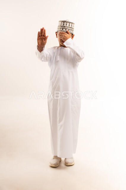Command to stop doing something, expressions of refusal and abstention, portrait of an Arab Gulf Omani boy wearing a dishdasha and a turban, putting his hand on his face and making gestures of prohibition and feeling angry and upset, full body image, white background