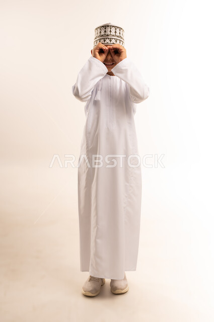 محاولة رؤية الأشياء البعيدة، حب الاستكشاف والمغامرة، التخطيط للأهداف والطموحات المستقبلية، بورتريه لولد عربي خليجي عماني يرتدي الدشداشة والكمة يقف باستقامة ويضع يديه على عينيه على شكل منظار ويقوم بالنظر، الطول الكامل للجسم، خلفية بيضاء