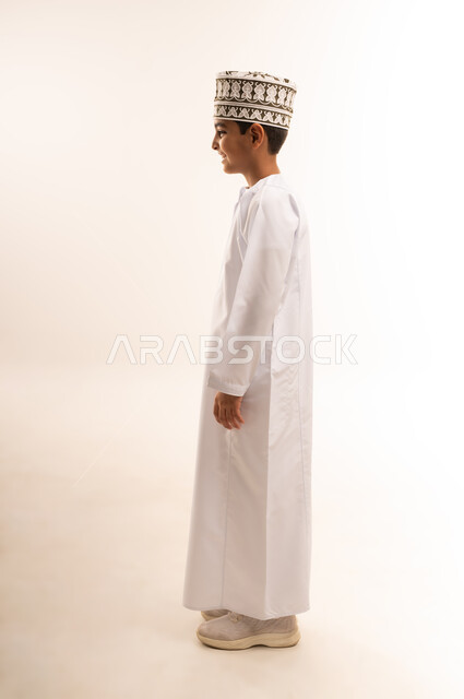 الاهتمام بالاناقه بالمظهر الخارجي، مفهوم الثقه بالنفس، بورتريه من الجانب لولد عربي خليجي عماني يرتدي الدشداشة والكمة يقف باستقامة، صورة الجسم بالطول الكامل، خلفية بيضاء