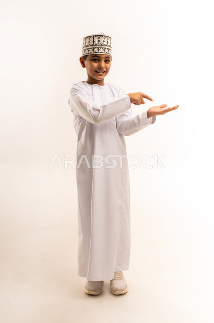 الامساك بعنصر بإيماءات الاعجاب، مفهوم النقر باستخدام حاسة اللمس, بورتريه لولد عربي خليجي عماني يرتدي الدشداشة والكمه العمانية يقف باستقامة يرفع يده ويقوم بالاشارة باصبع السبابة الى شيء ما, صورة الجسم بالطول الكامل، خلفية بيضاء