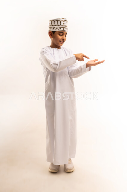 مفهوم النقر باستخدام حاسة اللمس, بورتريه لولد عربي خليجي عماني يرتدي الدشداشة والكمه العمانية يقف باستقامة يرفع يده ويقوم بالاشارة باصبع السبابة الى شيء ما, الامساك بعنصر بإيماءات الاعجاب، صورة الجسم بالطول الكامل، خلفية بيضاء