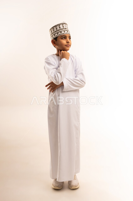 Focus, absent-mindedness and contemplation, finding a solution to a problem, contemplation and planning for the future, portrait of an Arab Gulf Omani boy wearing a dishdasha and a kummah, standing straight, putting his hand on his chin and looking up with thinking gestures, full-length body image, white background
